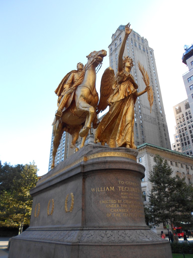 A gold statue of a man on a house. A female angel is in front with arm raised.