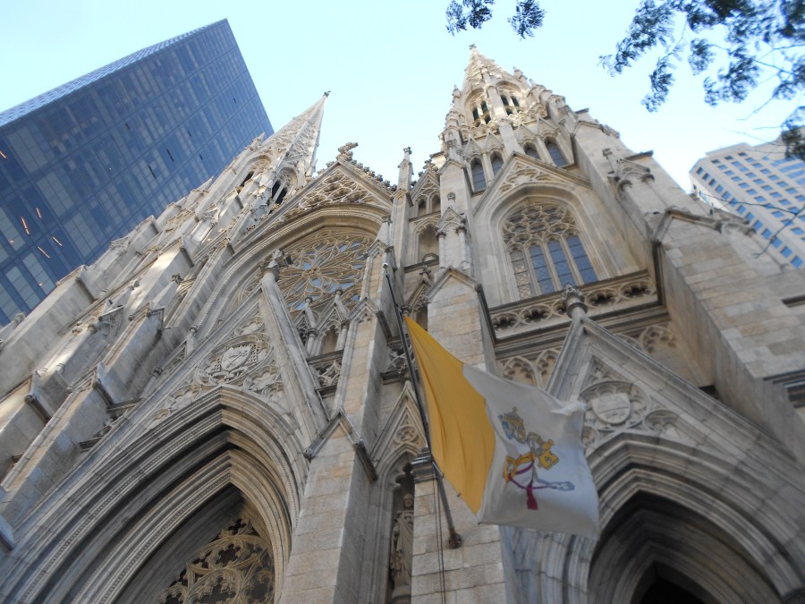 Looking up a St Patrick's Cathedral in New York City. Papal flag is flying. Modern skyscraper is next door.