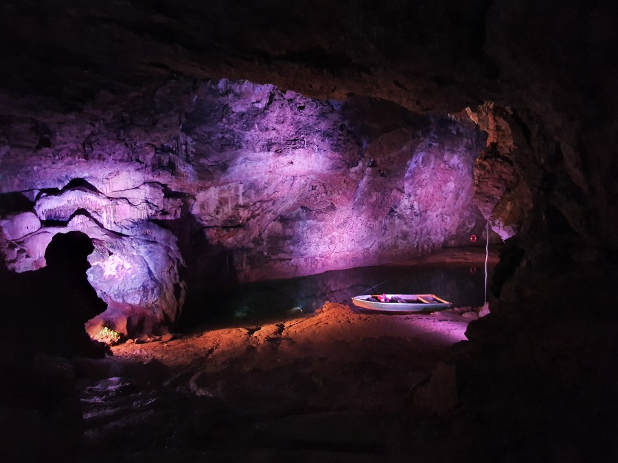 A cave lit purple, a small boat is on the end of a calm pool of water. A rock said to resemble a witches head is in shadow on the left
